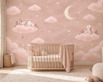 Puede incluir: Una habitación infantil con una cuna ovalada de madera clara y una manta beige, frente a un mural de pared con un cielo rosa empolvado, nubes de acuarela, estrellas doradas, una luna creciente y dos conejos blancos sobre nubes rosas.