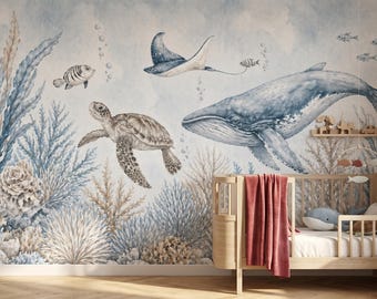 Puede incluir: Un mural de pared con una escena oceánica en acuarela que muestra una gran ballena azul, una tortuga marina, una mantarraya y varios peces nadando entre corales y algas, situado detrás de una cuna de madera con una manta roja y un peluche.