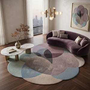 Peut inclure: Un grand tapis circulaire présentant des formes arrondies superposées dans des tons sourds de rose poudré, vert sauge, bleu ardoise et crème. Le tapis est posé sur un parquet en bois foncé dans une pièce avec un canapé violet incurvé.