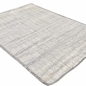 Peut inclure: Un tapis rectangulaire aux tons neutres, avec un motif tissé en forme de grille. Le tapis présente une palette de couleurs gris clair et blanc cassé, avec des bordures gris plus foncées. La texture semble légèrement en relief.