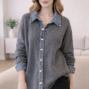May include: A gray button-down shirt with a denim collar and cuffs. The shirt has gold-colored buttons and is worn with black shorts. The model is wearing a gold necklace.