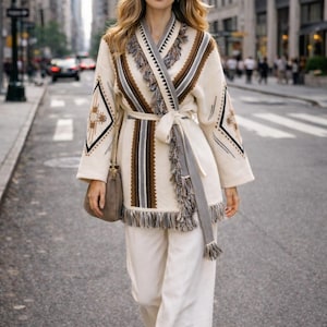 Può includere: Una donna indossa una giacca avvolgente color crema con motivi geometrici marroni, neri e grigi, abbinata a pantaloni bianchi. La giacca ha frange e una cintura abbinata. Una piccola borsa a tracolla grigia completa il look.