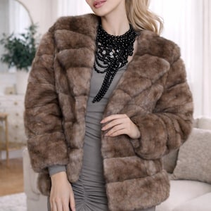 Puede incluir: Un abrigo de piel sintética marrón con un diseño clásico. El abrigo tiene un cuello ancho y mangas largas. La modelo lleva un vestido gris y un collar de cuentas negro. El abrigo es una opción de abrigo elegante.