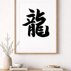 May include: Black ink calligraphy art print of the Japanese kanji character for "dragon" on white paper, framed in a light wood frame. The artwork is displayed on a wooden shelf with decorative items.