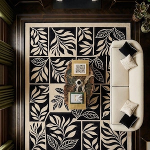 May include: Overhead view of a living room featuring a large black and cream-colored rug with a leaf pattern. A cream-colored sofa with black and cream pillows sits on the rug. A wooden coffee table with framed art and a cup of coffee is in the center.