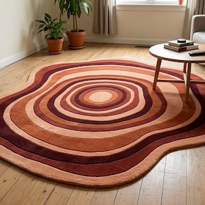 Puede incluir: Una alfombra de forma orgánica con círculos concéntricos en tonos marrones, óxido y melocotón. La alfombra, de aproximadamente 180 cm por 150 cm, está colocada sobre un suelo de madera, añadiendo calidez y un ambiente retro al salón.