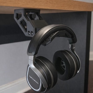 May include: Black and silver Turtle Beach headphones suspended from a black under-desk headphone mount. The headphones feature large earcups and a silver band. The mount is fixed to a wooden desk.