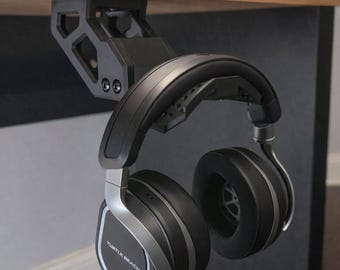 May include: Black and silver Turtle Beach headphones suspended from a black under-desk headphone mount. The headphones feature large earcups and a silver band. The mount is fixed to a wooden desk.