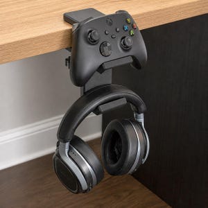 May include: Black desk-mounted gaming headset and controller holder. The holder is attached to a wooden desk and holds a black Xbox controller and a pair of black and silver headphones. The holder is made of black plastic.