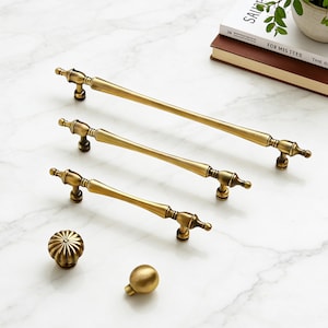 May include: A collection of antique brass cabinet hardware, including three different-sized pull handles and two knobs. The handles have a classic design with decorative end pieces. The knobs include a round, ribbed knob and a smooth, spherical knob.
