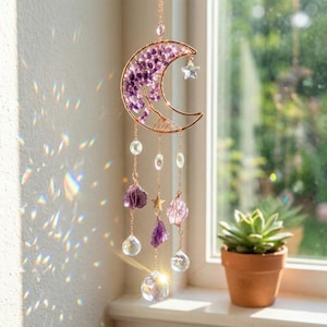 Puede incluir: Un atrapasoles en forma de luna de color cobre con una estrella y cristales colgantes. La luna está llena de piedras de amatista moradas. Cuentas de cristal transparente y trozos de amatista cuelgan de la luna. Una pequeña suculenta en maceta está en el fondo.