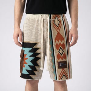 Könnte beinhalten: Beige Strickshorts mit Kordelzugbund und gemusterten Einsätzen. Die Einsätze zeigen geometrische Designs in Schwarz, Türkis, Orange und Braun. Auf der linken Seite ist eine kleine braune Tasche zu sehen.