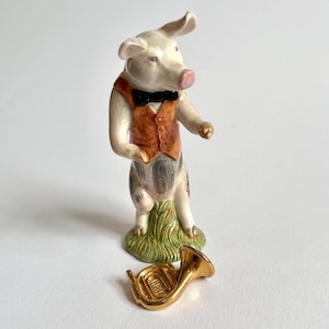 May include: A ceramic pig figurine wearing a brown waistcoat and a black bow tie, standing on a patch of green grass. A small, gold-coloured French horn lies nearby. The pig has a pink snout and is painted with detailed features.