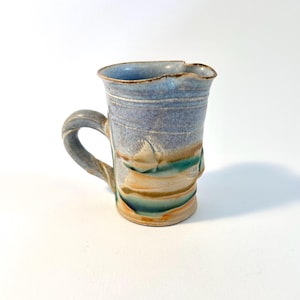 May include: Handmade ceramic mug with a unique, textured design. The mug features a blue and grey colour scheme with bands of tan and teal. It has a twisted handle and a slightly irregular rim, showcasing its artisanal quality.