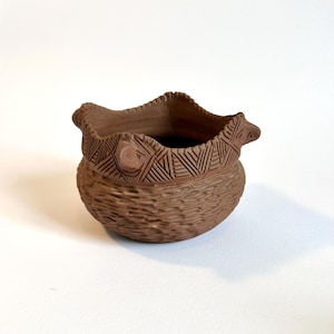 May include: A small, brown ceramic pot with a textured, woven-like body. The rim has a decorative geometric pattern and small, raised details. The pot is likely used for decorative purposes or as a small planter.