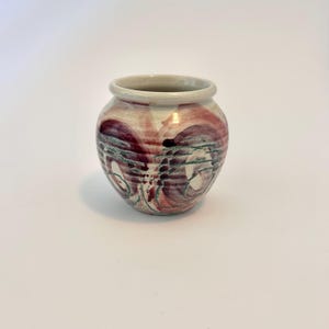 May include: A small, round ceramic vase with a wide opening. The vase has a cream-coloured rim and a body decorated with swirling patterns in shades of burgundy, purple, and green. The vase is a decorative item.