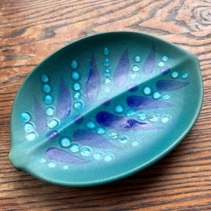 May include: A leaf-shaped ceramic dish with a matte teal finish. The surface features a decorative pattern of dark blue painted leaf shapes and raised, glossy light blue circular dots arranged in lines radiating from the centre vein.