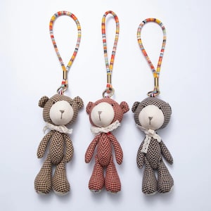 May include: Three teddy bear keychains with plaid patterns in brown, red, and gray. Each bear has a white face, a small lace collar, and a colorful woven strap. The keychains are hanging against a white background.