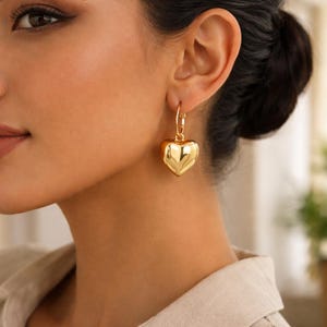 May include: A close-up of a gold heart-shaped earring. The earring is a hoop style with a large, puffy heart pendant. The gold is shiny and reflective. The earring is worn on a person's ear.