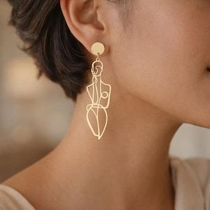May include: Gold-colored dangle earrings featuring a minimalist outline of a person. The earrings have a circular stud that attaches to the ear, with a delicate, linear design of a figure suspended below. The design is a continuous line, creating a modern, artistic look.