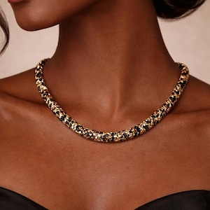 May include: A gold-toned necklace with black accents. The necklace is a choker style, featuring a woven design with alternating gold and black elements. The jewelry is worn around the neck, against a dark skin tone, and a black top.