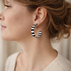 May include: Black and white striped hoop earrings. The earrings are a bold, geometric design, featuring alternating black and white stripes. The earrings are worn on the ear, showcasing the modern, graphic pattern. The earrings are a stylish accessory.