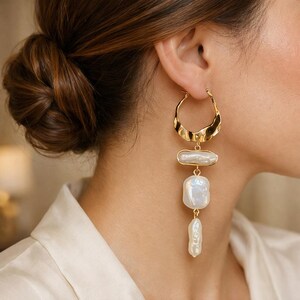 May include: Gold-tone hoop earrings with a textured design and three dangling, irregularly shaped, white pearl drops. The earrings are worn on a person's ear, with the hair pulled back in a bun.