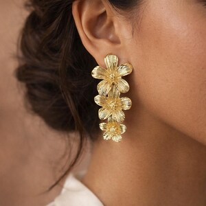 May include: Gold-colored flower earrings. Each earring features three textured, cascading flower shapes. The earrings are worn on the ear, with the top of the earring near the ear and the bottom of the earring hanging down.