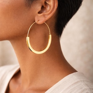 Könnte beinhalten: Goldfarbene Creolen. Die Ohrringe haben ein abgerundetes, offenes Design mit einem flachen, rechteckigen Boden. Die Ohrringe sind hell und glänzend goldfarben und werden am Ohr getragen.