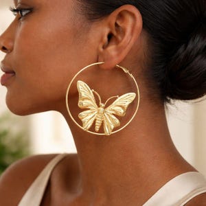 May include: A large gold-toned hoop earring featuring a central three-dimensional butterfly charm. The butterfly has detailed, textured wings and is positioned within the circular frame of the hoop, creating a bold and metallic accessory piece.