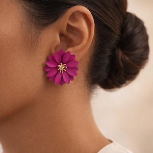 May include: A close-up of a person's ear wearing a magenta flower-shaped earring. The earring has multiple petals and a gold-colored center. The background is a blurred neutral tone.