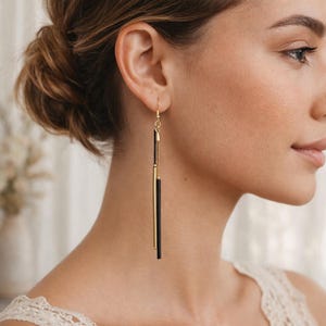 May include: Gold-tone drop earrings with a sleek, modern design. Each earring features a slender black bar suspended from a gold chain and a gold cylindrical accent. The earrings are shown on a person's ear.