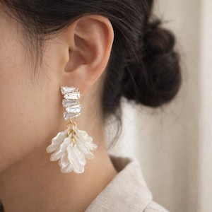 May include: Close-up of a white and gold earring. The earring features a cluster of rectangular, clear crystals and a dangling, flower-shaped design with white, petal-like elements. The earring is attached to a gold-colored hook.