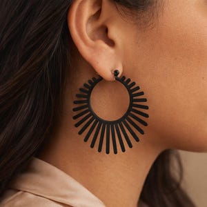 May include: Black hoop earrings with a sunburst design. The earrings feature a circular shape with radiating lines, creating a bold and geometric look. The earrings are a dark matte black color, offering a modern and stylish accessory.