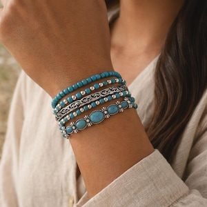 May include: A collection of turquoise and silver bracelets stacked on a wrist. The bracelets include beaded, engraved, and oval turquoise stone designs. The jewelry is displayed against a neutral background, highlighting the vibrant blue of the stones.