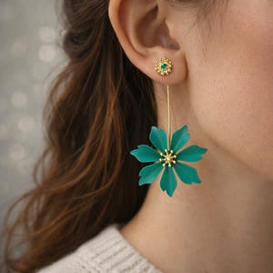 May include: A pair of gold-toned earrings. One earring is a small stud with a green center stone. The other earring is a long drop with a teal flower design and a gold-toned center.
