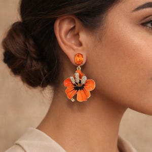 May include: Orange flower earrings with a gold-tone metal finish. The earrings feature a large, enamelled flower design with a cluster of small, clear crystals in the centre. An oval-shaped orange gemstone tops each earring.