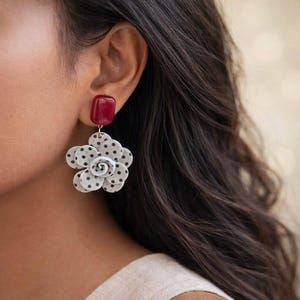 May include: A close-up of a floral earring. The earring features a dark red square stud, connected to a white flower with black polka dots. The flower has a white spiral center. The earring is hanging from a gold-colored hook.