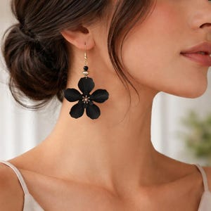 Op de afbeelding: Een close-up van een vrouw die een paar zwarte bloemenoorbellen draagt. Elke oorbel heeft een grote, vijfbladige bloem met een klein, sprankelend centrum. De oorbellen hangen aan goudkleurige haken.