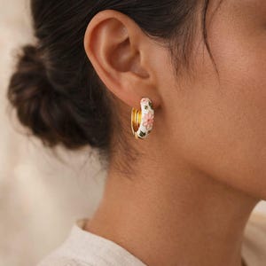 May include: Gold hoop earrings with a white enamel finish, adorned with pink and green floral designs. The earrings are a classic hoop style, featuring a delicate and feminine aesthetic.