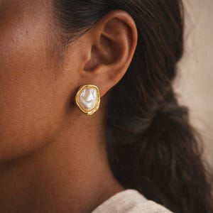May include: Gold-tone stud earrings with a large, irregular-shaped white pearl center. The pearl is surrounded by a textured gold border. The earrings are shown on an ear, suggesting a jewelry product.