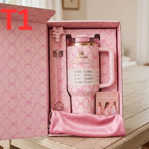 May include: A pink Stanley tumbler with a bow pattern, a matching straw with a bow topper, and a pink velvet pouch inside a gift box. The tumbler features the Stanley and LoveShackFancy logos, a handle, and a product information label on the front.