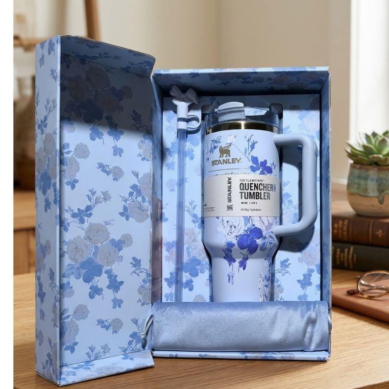 K&ouml;nnte beinhalten: Ein hellblauer Stanley Quencher-Becher mit Griff, Strohhalm und Blumendesign, pr&auml;sentiert in einer passenden Geschenkbox. Die Box und der Becher weisen ein blau-wei&szlig;es Blumenmuster auf. Der Becher tr&auml;gt den Text "QUENCHER TUMBLER".