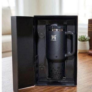 May include: A black Stanley travel mug with a handle, presented in a black box. The mug features the Stanley logo and a floral design at the base. The box is open, revealing the mug's interior.