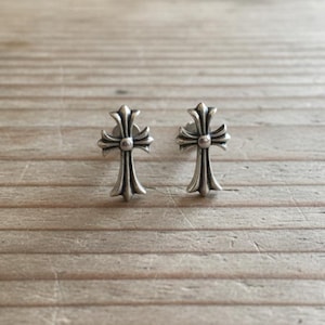 May include: A pair of silver-colored cross stud earrings. Each earring features a detailed cross design with a central sphere and ornate arms. The earrings are displayed on a light brown wooden surface.