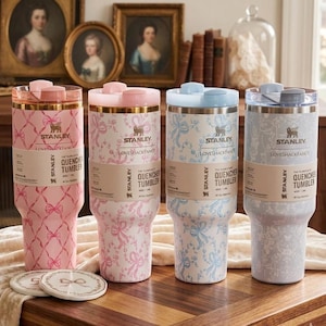 May include: Four Stanley Quencher tumblers, 1.18 litres, featuring LoveShackFancy patterns. Two are pink with ribbon designs, one is light blue with floral patterns, and one is grey-blue with floral patterns. Each has a lid and a branded paper band.