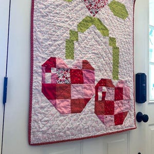 May include: A handmade quilt featuring a floral design with a heart-shaped flower and two heart-shaped fruits. The quilt has a white background with a diamond pattern, red polka dot border, and green and red accents.
