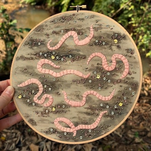 May include: A circular embroidery hoop with a brown background and pink embroidered worms. The background is decorated with small, colorful beads.