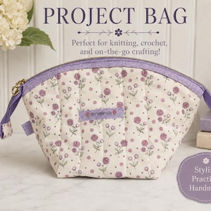 May include: A cream-coloured project bag with a floral pattern in purple. The bag has a purple zip and handles, with the text "PROJECT BAG" at the top. It is designed for knitting, crochet, and on-the-go crafting. The bag is handmade.