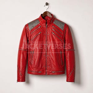 Puede incluir: Chaqueta de cuero roja con cuello alzado y múltiples bolsillos con cremallera. Los hombros presentan paneles grises. La chaqueta cuelga de una percha de madera sobre un fondo blanco.